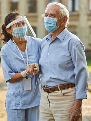 Nurse assisting elderly man outdoors, both wearing masks and protective gear.