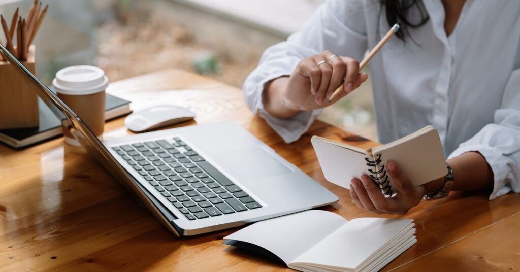 Person in white shirt taking notes at wooden desk with laptop.