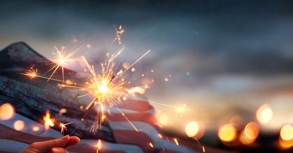 A hand holding a sparkler with a blurred American flag background.