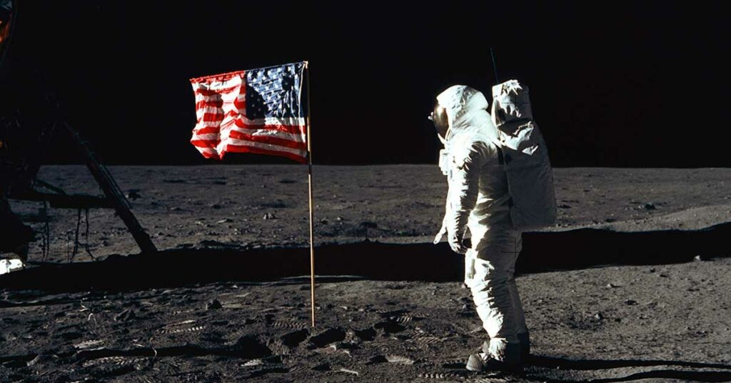Astronaut in a spacesuit stands beside the American flag on the lunar surface.