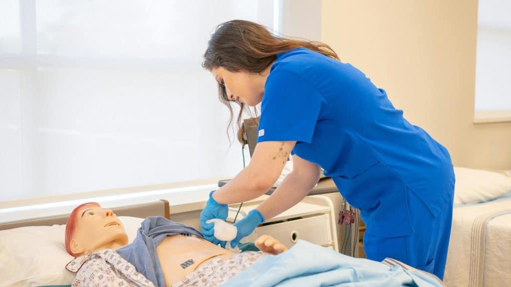 A healthcare worker in blue scrubs examines a patient lying on a medical bed in a hospital room.
