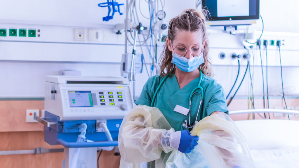 A nurse putting on PPE gear next to a bed and hospital equipment
