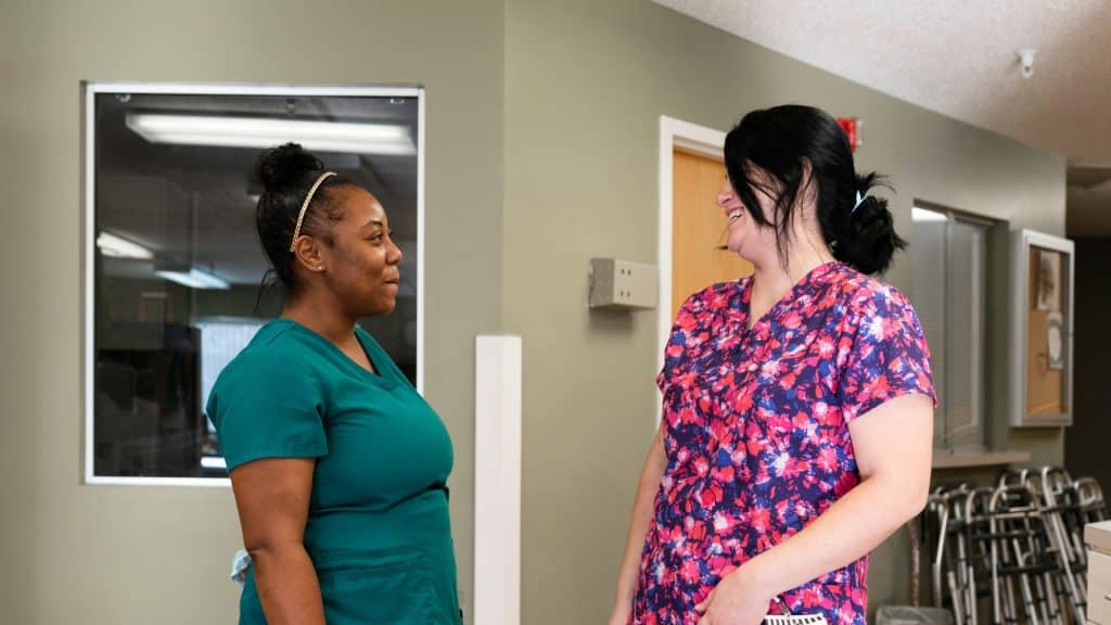 Two healthcare workers in scrubs are standing and talking in a hallway. One is wearing a teal scrub top, and the other is wearing a colorful scrub top.